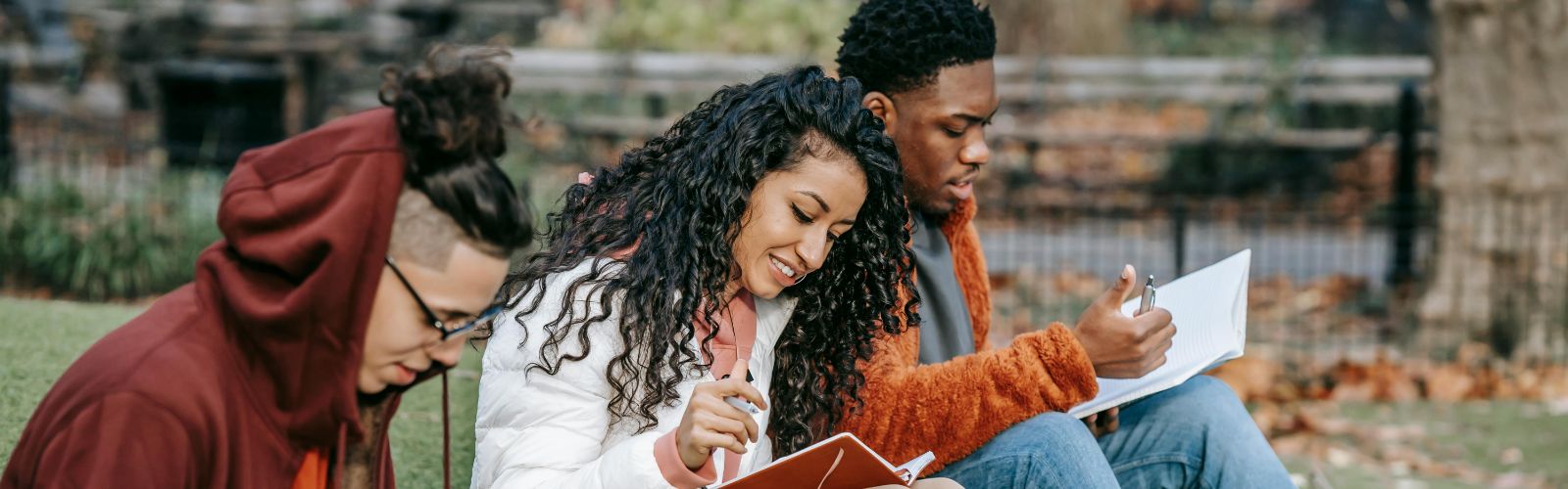 Three students are studying together outdoors, enjoying a collaborative learning experience in a relaxed park setting. The central figure is smiling, reflecting the positive aspects of group study.